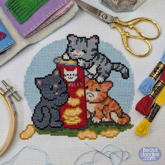 Cross-stitched design of three cats around a container of crisps. Embroidery tools surround the stitched area on white fabric.