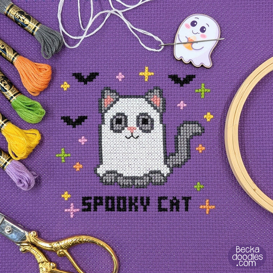 A cross-stitch design of a cat dressed as a ghost surrounded by bats and colorful stars and 'SPOOKY CAT' stitched below on purple Aida cloth. Surrounding the design are thread skeins, a wooden hoop, scissors, and a ghost needle minder.