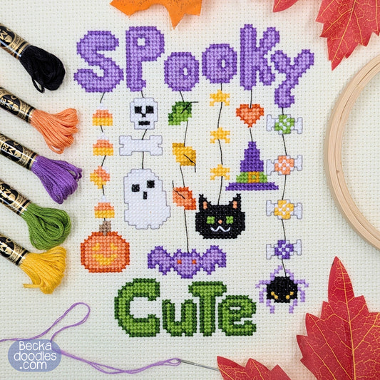 Cross-stitch design on cream Aida fabric, featuring the words 'Spooky Cute' in purple and green stitched letters, with Halloween motifs including ghosts, pumpkins, a black cat, stars, leaves, and cand corn. Surrounded by sewing supplies.