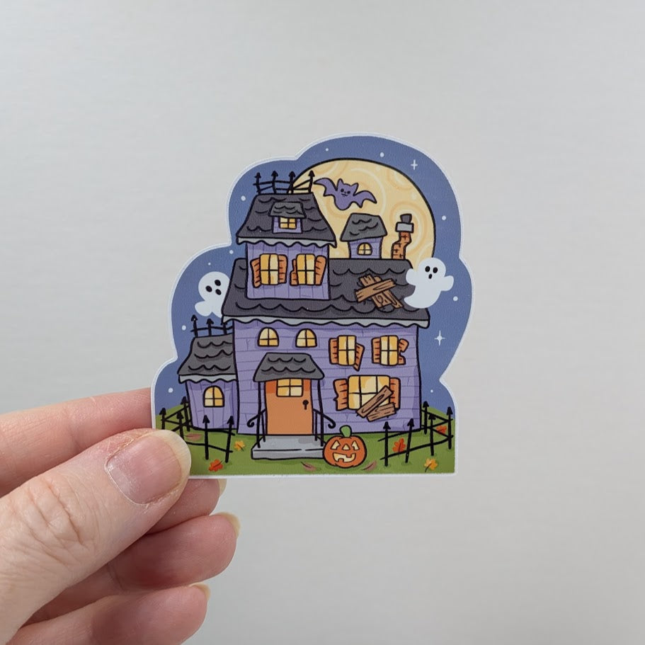 Halloween-themed sticker of a haunted house held by a hand on a light gray background