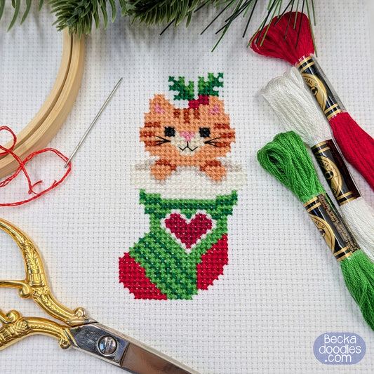 Cross-stitched cat in a stocking with embroidery supplies on white fabric background