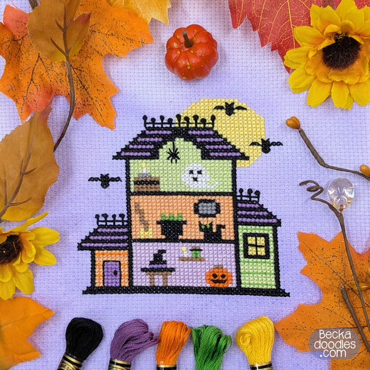 Cross-stitch design of a tiny haunted house with Halloween filled rooms, stitched on dyed lavender Aida cloth and surrounded by autumn leaves, sunflowers, a small pumpkin, and embroidery floss skeins. 