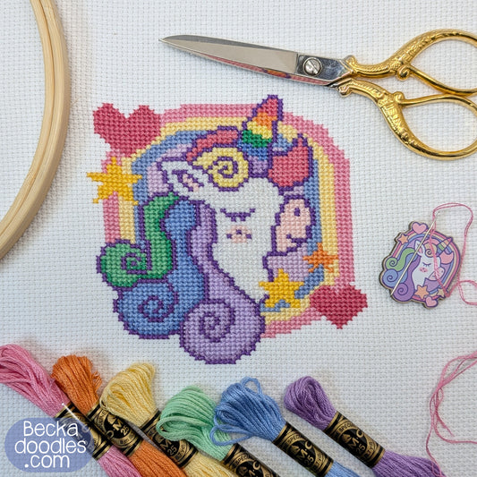 Cross-stitched unicorn design with embroidery threads and scissors on a white background