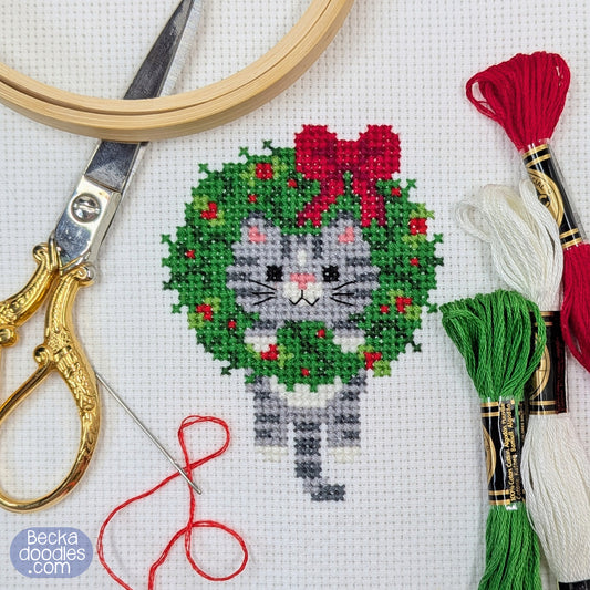 Cross-stitched gray cat in a wreath with embroidery supplies on  white Adia cloth.